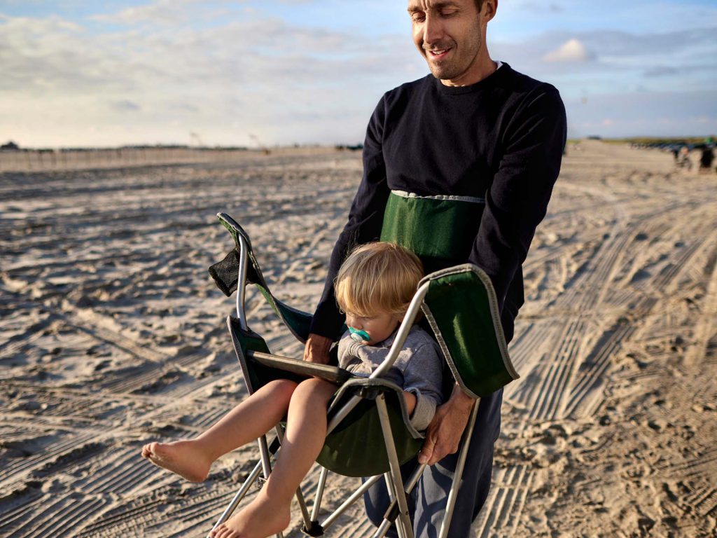 Mann trägt Kind im Strandstuhl über den Strand. Thema Herzinfarkt