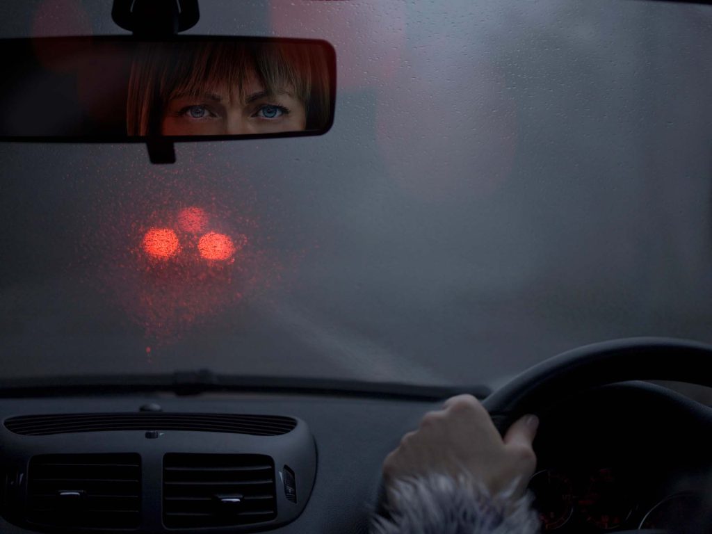 Junge Frau am Steuer des Autos bei Regen und blendenden Lichtern vor ihr. Migräne Kolumne