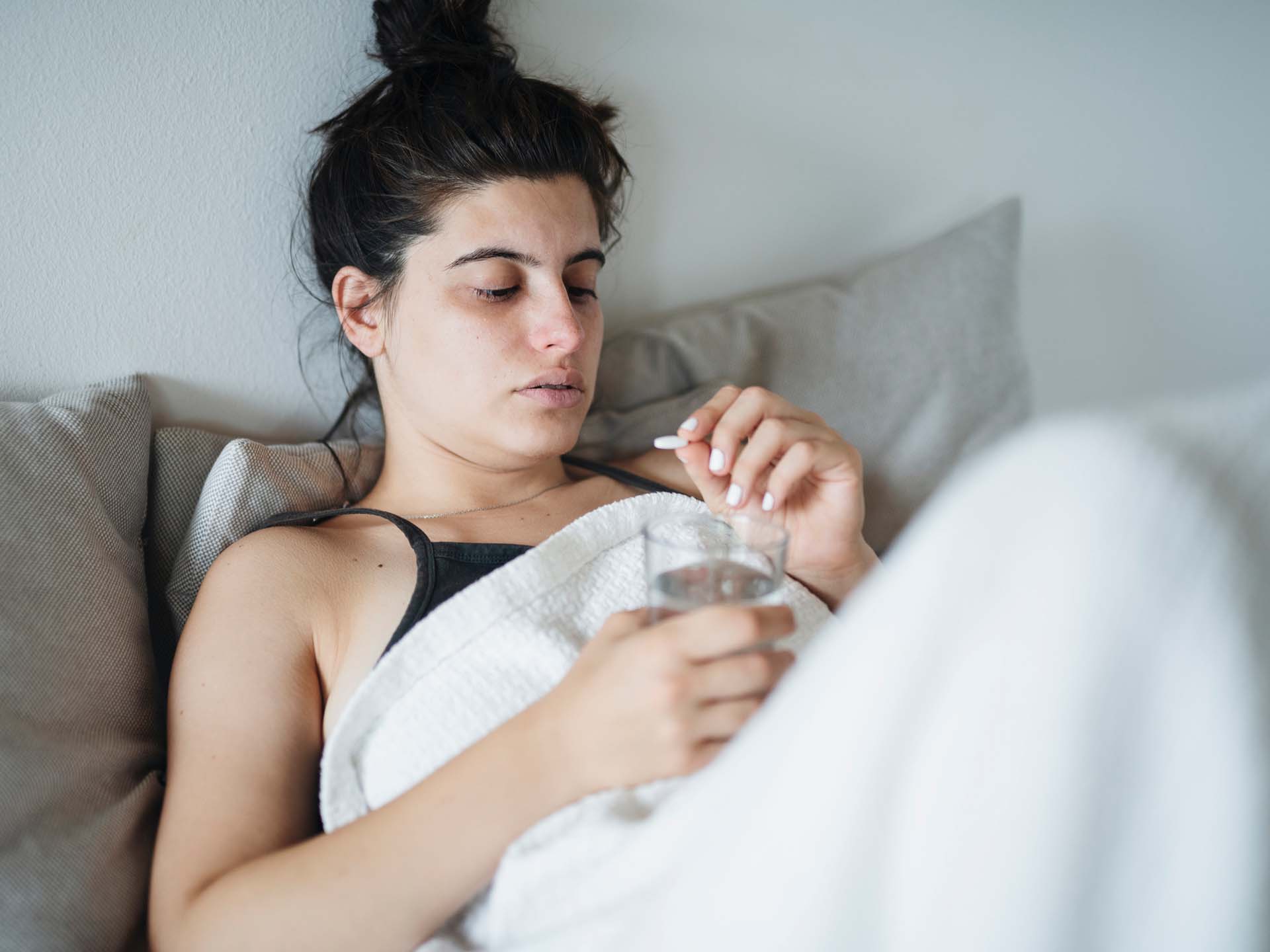 Junge Frau mit Kopfschmerzen sitzt im Bett und schaut eine Tablette an. Thema chronische Migräne