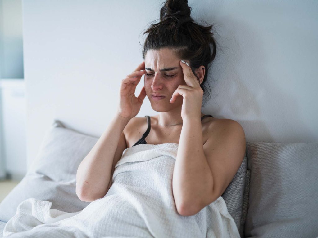 Junge Frau mit Kopfschmerzen sitzt im Bett. Thema chronische Migräne