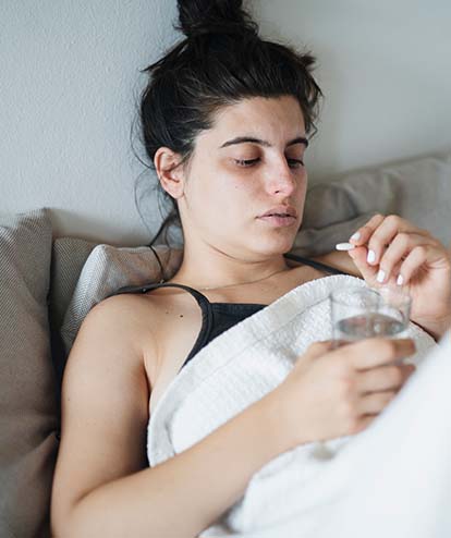 Junge Frau mit Kopfschmerzen sitzt im Bett und schaut eine Tablette an. Thema chronische Migräne